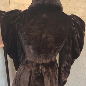 May include: A black velvet jacket with a high collar and dramatic, puffed sleeves. The jacket has a fitted waist and a flared peplum. The fabric appears soft and luxurious, with a slight sheen. The back of the jacket is visible.