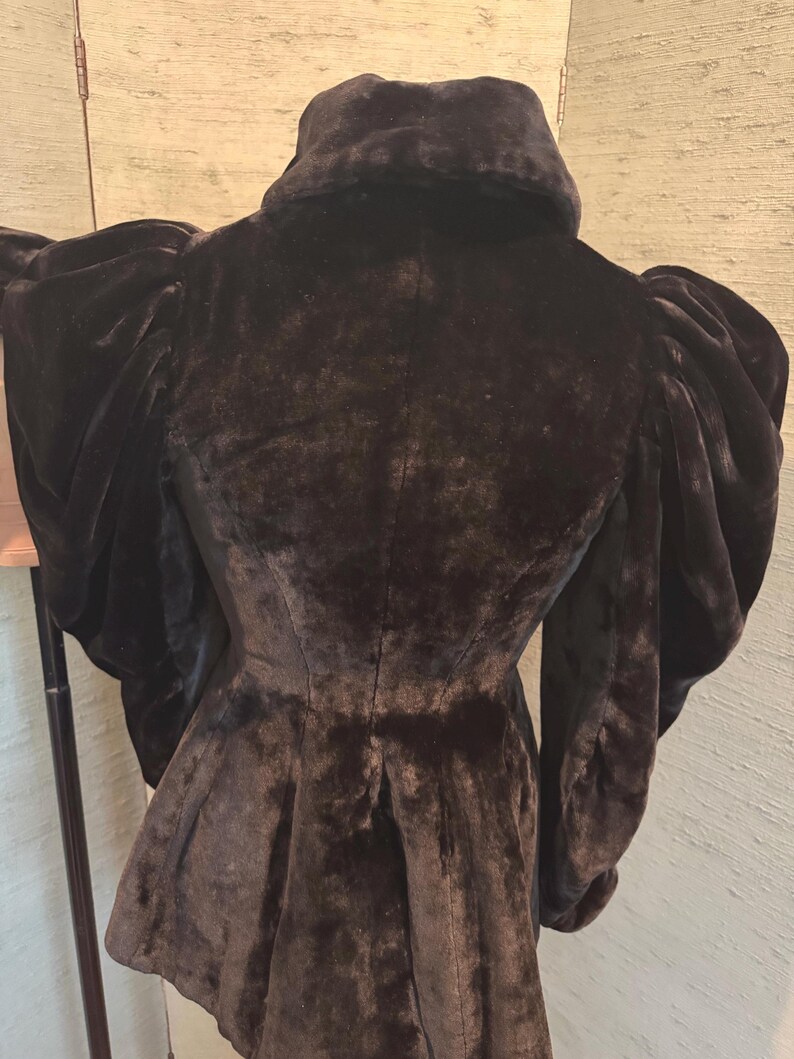 May include: A black velvet jacket with a high collar and dramatic, puffed sleeves. The jacket has a fitted waist and a flared peplum. The fabric appears soft and luxurious, with a slight sheen. The back of the jacket is visible.