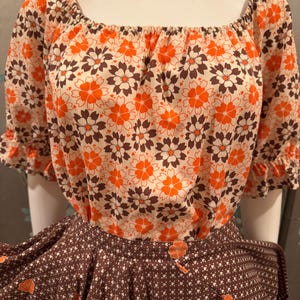 May include: A vintage-style two-piece outfit featuring a floral print. The top is off-the-shoulder with orange, brown, and white flowers on a cream background. The skirt is brown with a white heart and orange flower pattern.