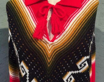 May include: A colorful poncho with a red collar and tassel tie. The poncho features a geometric pattern in black, white, gold, and green. The garment is displayed on a mannequin.