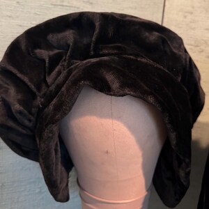 May include: A black velvet bonnet is displayed on a mannequin head. The bonnet has a soft, textured appearance and is gathered at the top. The dark color and plush material suggest a vintage or elegant style. The background is a light green and brown.