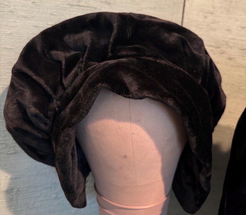 May include: A black velvet bonnet is displayed on a mannequin head. The bonnet has a soft, textured appearance and is gathered at the top. The dark color and plush material suggest a vintage or elegant style. The background is a light green and brown.