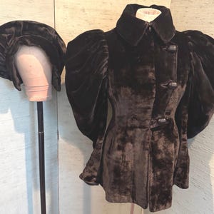 May include: A dark brown velvet jacket with large, puffed sleeves and a matching hat. The jacket has a high collar and two large buttons. The hat is round and has a wide brim. The jacket and hat are displayed on a mannequin.