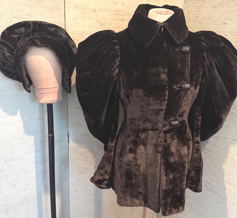 May include: A dark brown velvet jacket with large, puffed sleeves and a matching hat. The jacket has a high collar and two large buttons. The hat is round and has a wide brim. The jacket and hat are displayed on a mannequin.