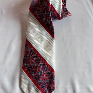 Puede incluir: Una corbata con un patrón geométrico repetitivo en rojo y azul oscuro, contrastado por bandas diagonales de tela color hueso con textura acanalada y finas rayas de color azul claro. La corbata tiene un extremo puntiagudo y una parte superior doblada.