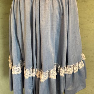 May include: A light blue skirt featuring a subtle swiss dot texture and an elasticated waistband. The garment includes a tiered design with a white lace trim accentuating the horizontal seam near the bottom hem, creating a gathered silhouette.