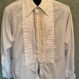 Pode incluir: Uma camisa social branca de manga comprida com um painel frontal plissado e um detalhe central de babados verticais. A camisa tem gola pontiaguda, punhos abotoados com bordas de babados e uma etiqueta visível na parte inferior frontal.
