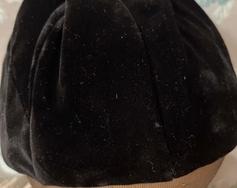 Puede incluir: Un sombrero de terciopelo negro con una corona redonda y un ala ancha. El sombrero está hecho de terciopelo suave y afelpado y tiene una textura lisa y uniforme.