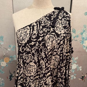 May include: Black and cream-colored one-shoulder dress with a floral pattern. The dress is displayed on a mannequin. The fabric has a repeating design of swirling floral and leaf motifs. A black tie detail accents the shoulder.