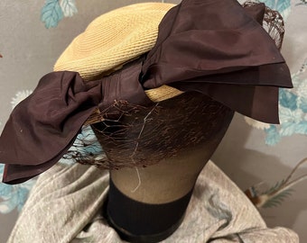May include: A straw hat with a large brown bow and a brown net veil. The hat is on a mannequin head.