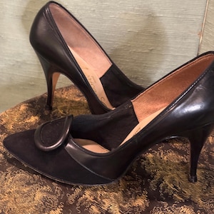 Può includere: Un paio di scarpe con tacco alto in pelle nera con punta a punta e una fibbia decorativa sul davanti. Le scarpe sono realizzate in una combinazione di pelle e camoscio.
