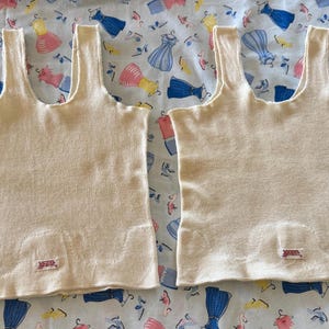 May include: Two sleeveless cream-colored knit tank tops with wide shoulder straps and a scoop neckline. Each garment features a small rectangular white label with red text near the bottom hem, displaying the brand name Vanta.