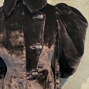 May include: A close-up of a black velvet coat with a large collar and three large buttons. The coat has puffed sleeves and a tailored fit, suggesting a vintage or retro style.