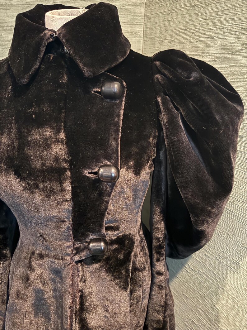 May include: A close-up of a black velvet coat with a large collar and three large buttons. The coat has puffed sleeves and a tailored fit, suggesting a vintage or retro style.
