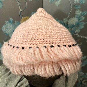 Op de afbeelding: Een roze gebreide muts met een franje rand en zwarte kralen rond de rand.