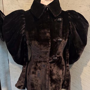 May include: A black velvet coat with a wide collar and large, puffed sleeves. The coat features a button-down front and a tailored silhouette. The fabric has a soft, textured appearance, suggesting a luxurious feel. The coat is displayed on a mannequin.