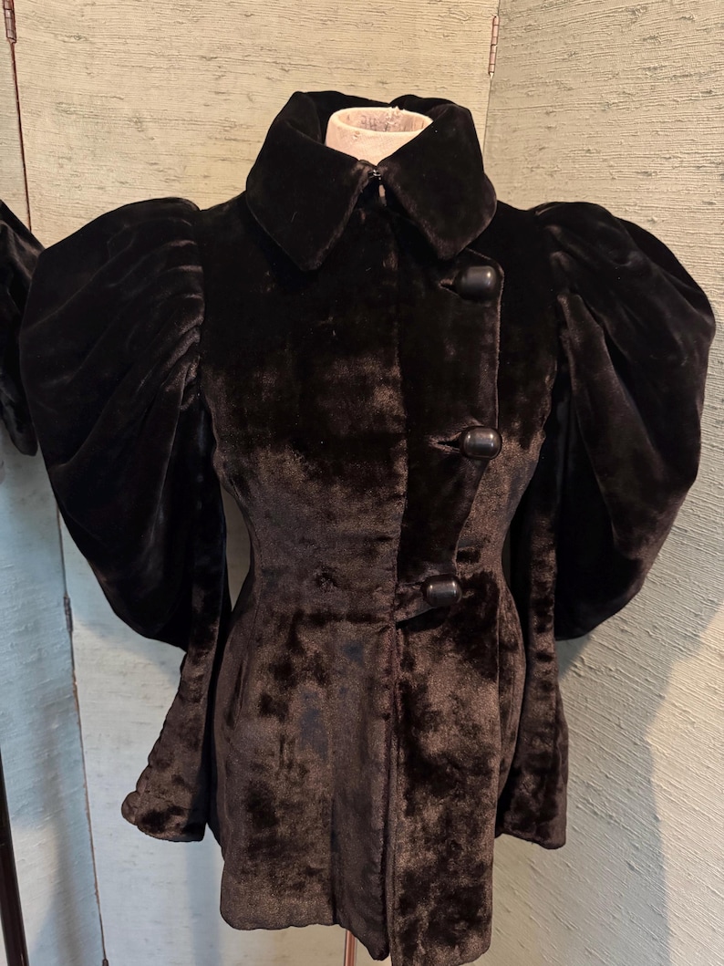 May include: A black velvet coat with a wide collar and large, puffed sleeves. The coat features a button-down front and a tailored silhouette. The fabric has a soft, textured appearance, suggesting a luxurious feel. The coat is displayed on a mannequin.