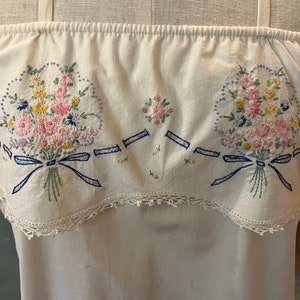 May include: White cotton dress with a delicate lace trim and embroidered floral bouquets with blue ribbons. The dress has a gathered neckline and a relaxed fit.
