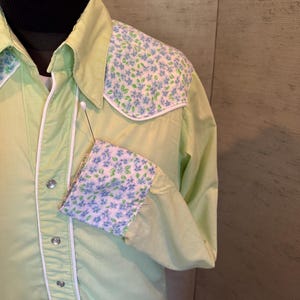 May include: A light green button-down shirt with floral patterned shoulder and cuff accents. The shirt features white piping and silver snap closures. The floral pattern includes blue and green flowers on a white background.