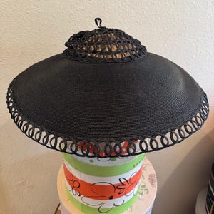May include: A black sun hat with a wide brim and a decorative trim. The hat features a woven crown and a looped trim around the edge of the brim. The hat is displayed on a colorful round box.