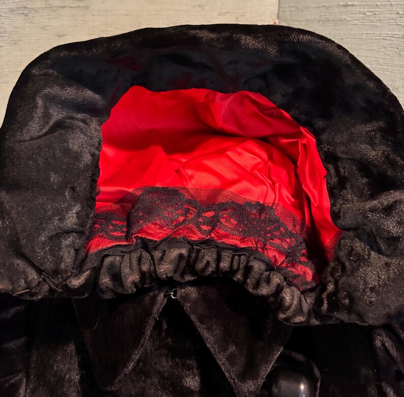 May include: Black hooded cloak with a red satin lining. The hood features a black lace trim. The cloak has a button closure.