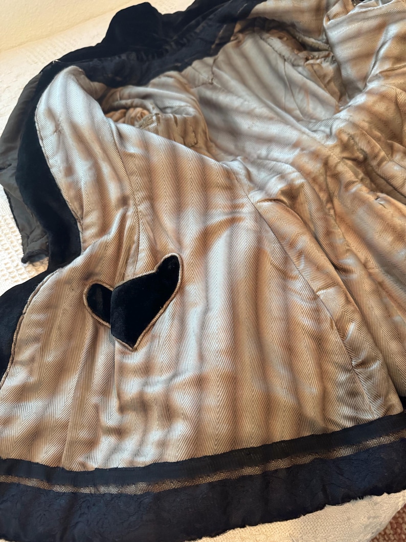May include: Black velvet coat with a light brown herringbone patterned lining. The coat features a heart-shaped detail and black lace trim. The interior lining is visible, showcasing the coat's construction and materials.