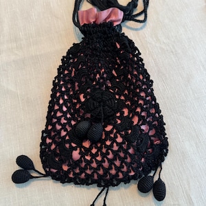 May include: A black crocheted drawstring bag with a pink satin lining and black tassel accents. The bag has a delicate floral pattern and is adorned with multiple black crocheted pom-poms.