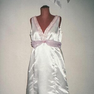 Puede incluir: Un vestido de satén blanco con escote en V y una cinta morada clara alrededor de la cintura. El vestido sin mangas tiene una falda fluida. Un lazo a juego es visible en la parte superior. El vestido se exhibe en un maniquí.
