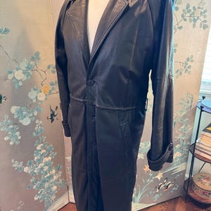 Könnte beinhalten: Ein langer, schwarzer Ledermantel mit Reverskragen und Knopfverschluss. Der Mantel hat ein klassisches Trenchcoat-Design mit einreihiger Front, langen Ärmeln und einer taillierten Passform. Der Mantel wird auf einer Schaufensterpuppe präsentiert.