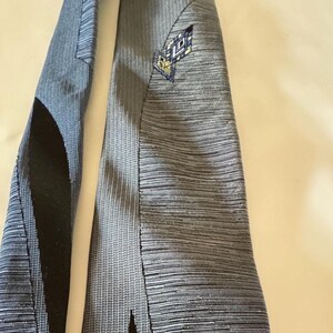 May include: A necktie featuring a textured grey fabric with horizontal lines. The tie includes a black panel on the left side and a small, embroidered geometric diamond-shaped emblem in blue, white, and yellow near the top right section.