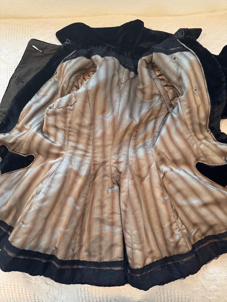 May include: An open black velvet coat with a light brown and tan striped lining. The coat has a notched collar and a black trim along the edges. The interior lining is visible, showing the coat's construction and details.