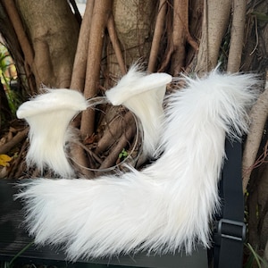 Puede incluir: Un conjunto de cosplay de piel sintética blanca con una diadema con orejas y una cola esponjosa. Las orejas están unidas a una diadema de plástico transparente. La cola tiene una correa negra con una hebilla.
