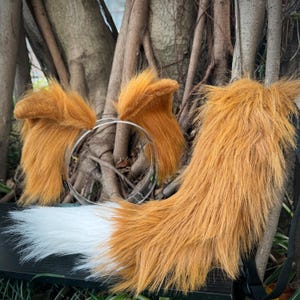 May include: A fox cosplay set with orange and brown faux fur ears on a clear headband, and a matching tail. The tail is predominantly orange and brown with a white tip, and has a black strap for attachment.