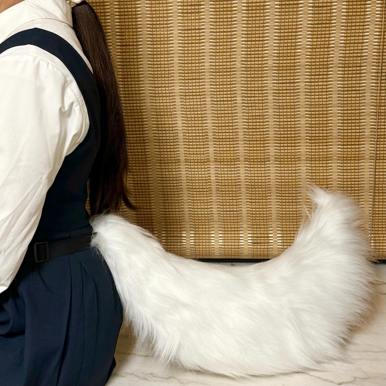 May include: A fluffy, white faux fur tail attached to a black belt. The tail is curved and has a soft, textured appearance. The background features a woven, light brown wall.