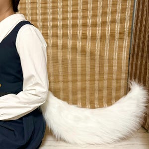 May include: A fluffy, white faux fur tail, curved and resting on a surface. The tail is thick and tapered, with a soft, textured appearance. The tail is a costume accessory.