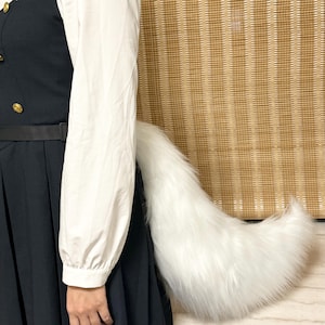 May include: A fluffy, white faux fur tail, likely for cosplay or costume use. The tail is large and tapered, with a full, soft appearance. The tail is attached to a black garment, with a white long-sleeved shirt visible.