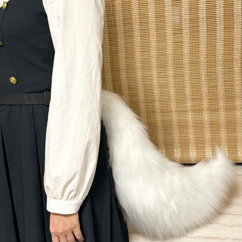 May include: A fluffy, white faux fur tail, likely for cosplay or costume use. The tail is large and tapered, with a full, soft appearance. The tail is attached to a black garment, with a white long-sleeved shirt visible.