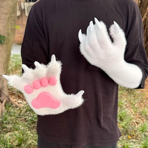 Puede incluir: Un par de guantes de piel sintética blanca con forma de patas de animales. Los guantes tienen largas garras blancas y almohadillas rosas. Los guantes están diseñados para ser usados en las manos.