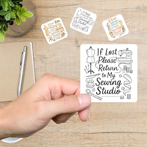 May include: A hand holding a white square sticker with the text "If Lost Please Return to My Sewing Studio" in black. The sticker features illustrations of sewing tools. Three smaller stickers with similar designs are also visible.
