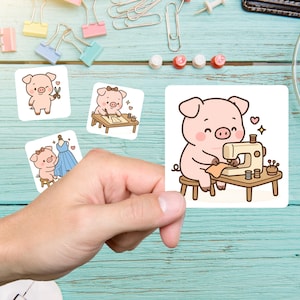 May include: A hand holding four square stickers with cartoon pig illustrations. One pig is sewing, another is drawing, a third is holding scissors, and the last is admiring a dress. The stickers are on a light blue wooden surface.