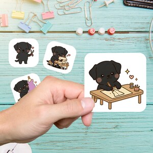 May include: A hand holding four square stickers featuring a black Labrador cartoon dog. The dog is depicted with sewing tools, a sewing machine, and drawing on a table. The stickers are on a light blue surface with office supplies.