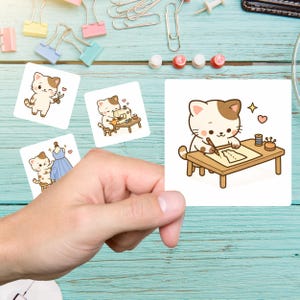 May include: A hand holding four square stickers featuring cartoon cats. The cats are depicted in various sewing-related activities, such as using scissors, sewing with a machine, designing a dress, and drawing on a table. The stickers have a white background.