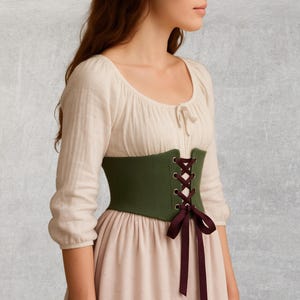 May include: A green corset with burgundy lacing and a matching bow. The corset is worn over a cream-colored blouse and a light pink dress. The outfit has a historical or renaissance style.