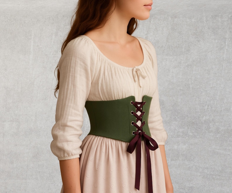 May include: A green corset with burgundy lacing and a matching bow. The corset is worn over a cream-colored blouse and a light pink dress. The outfit has a historical or renaissance style.