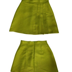 May include: A green mini skirt with a pleated front and a textured fabric. The skirt is shown from the front and back.