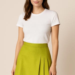 May include: A woman wearing a white t-shirt and a green pleated skirt. The skirt has a checkered pattern.