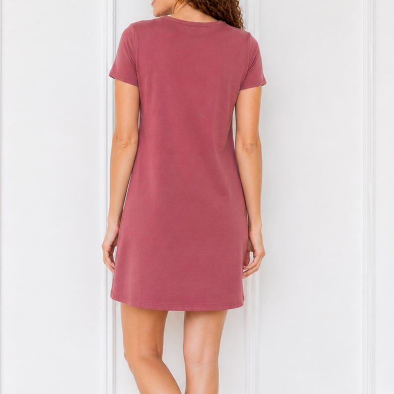 May include: A short-sleeved, solid mauve-colored dress featuring a simple round neckline and a straight, relaxed silhouette that falls above the knee. The garment is shown from the back, displaying a plain, unpatterned fabric construction.