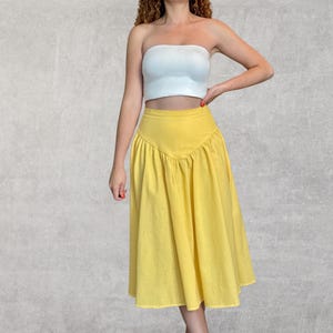 May include: A yellow midi skirt with a high waist and a flared silhouette. The skirt has a unique V-shaped design at the waist and is made of a lightweight fabric. The model is wearing a white tube top.