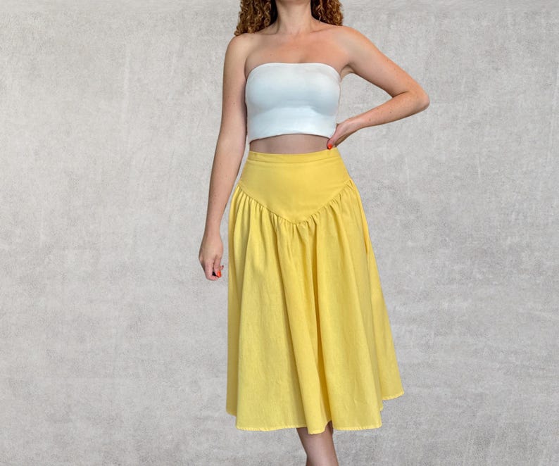 May include: A yellow midi skirt with a high waist and a flared silhouette. The skirt has a unique V-shaped design at the waist and is made of a lightweight fabric. The model is wearing a white tube top.