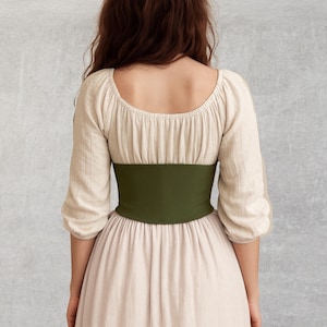 May include: A beige linen dress with a scoop neckline and three-quarter sleeves. A wide, olive green corset belt cinches the waist. The dress has a flowing skirt, creating a medieval or renaissance style.